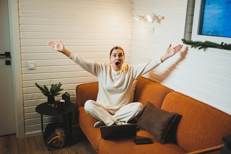 Excited woman on sofa with laptop, expressing surprise and joy in cosy living room setting