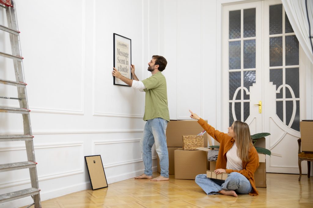 Couple decorating new home, hanging frame, surrounded by moving boxes in white living room